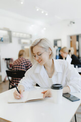 Student young woman writes content plan in notepad in cafe with coffee, white light