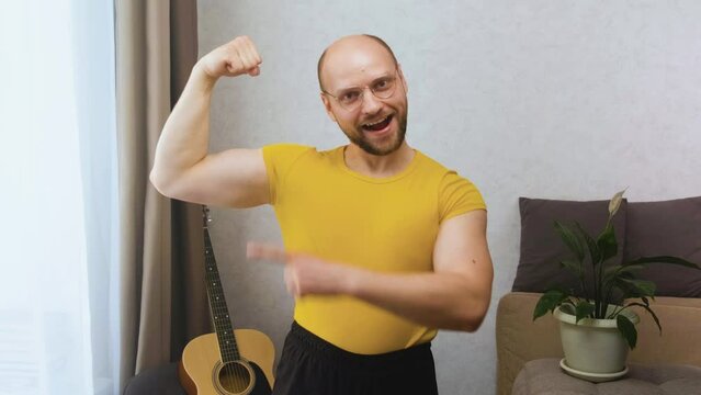 A funny athletic nerd man with glasses looks at the camera, smiles stupidly, shows a pumped-up bicep, points at it with his finger and moves his eyebrows