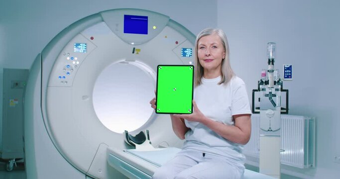 Mature woman is sitting at TC scanner bed. Female at room of MRI. Woman dressed up in white is showing screen of chroma key tablet. Female patient is posing at background of medical equipment.