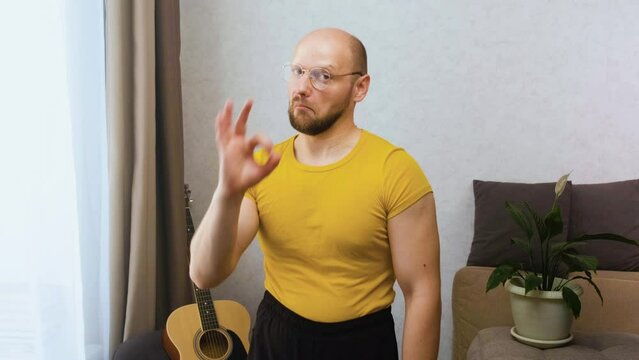 Funny bald and bearded nerd in glasses with high-stretched shorts muscles humorously nods approvingly and shows the OK gesture in the apartment at home