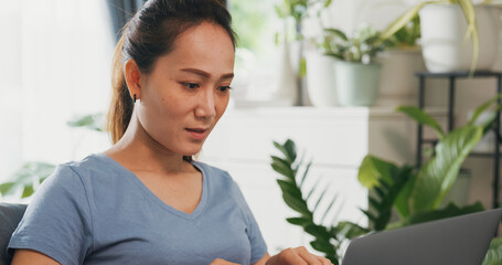 Young Asian woman wear casual calm sitting on wing chair use laptop computer thinking the idea for working full of plants in living room greenhouse. Coronavirus and stay home, Work from home concept.