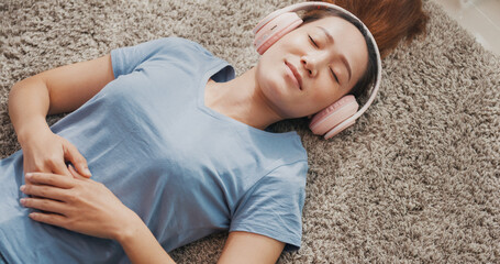 Young Asian university woman with casual wear headphone lay down on carpet floor listen music calm and relax take a rest in living room at home. Cozy home interior with indoor plants concept.