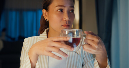 Close-up Young Asian businesswoman wear formal sit in front of desk focus on laptop typing finance report take rest drink cup of tea in cozy living room at home night. Overtime work concept.