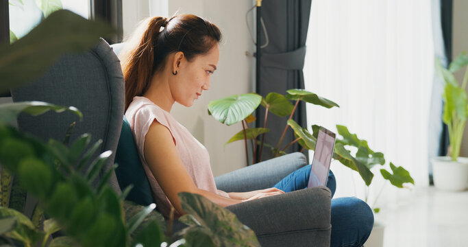 Young Asian woman wear casual sitting on wing chair use laptop computer focus on screen typing report feel tired stretching arm raise hand with plant in living room greenhouse. Work from home concept.