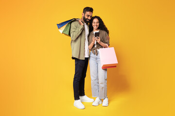 Middle Eastern Spouses Using Mobile Phone Shopping On Yellow Background
