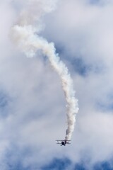 Vertical shot of a bi-plane stunt flying with a smoke trail in the blue sky with white clouds