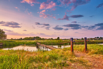 Landscape with sunset over river. Rural pedestrian old bridge. Evening with calm beautiful clouds on sky. Water reflection. Tranquil evening scene. Green filed and meadow. - 612479283