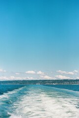 Vertical shot of a boat trail in the sea and the clear blue sky with landscape in the distance