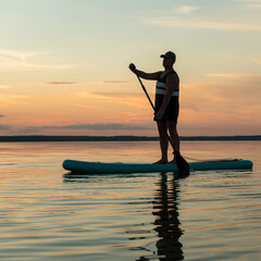 A man on a SUP board with a paddle swims in the calm water of the lake against the backdrop of the evening sky at sunset.