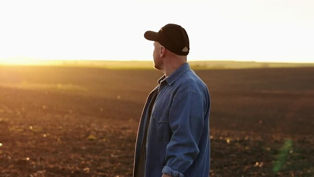 Side view of male farmer standing and looking in cultivated plowed field at sunset in spring. Owner agricultural farm is checking and examining farmland before sowing agriculture crops. Agribusiness