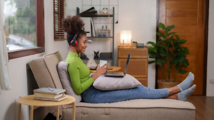 Obraz premium Young african american woman wearing headphones using laptop sitting on sofa in the living room and holding coffee, watching movie, listening to music, shopping online, watching live broadcast.