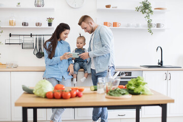 Happy young family of three in denim outfits looking at mobile screen behind kitchen table at home. Parents and kid searching for online recipe on phone with fresh ingredients for salad in foreground.