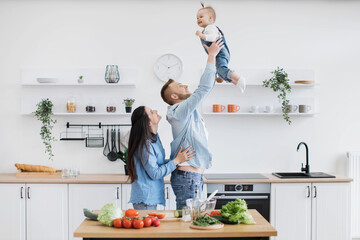 Handsome strong father raising child high in air while excited mother cheering her daughter up in dining room. Emotional couple and baby girl increasing happiness and fun during cooking meal at home.