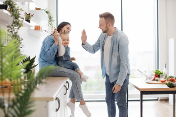 Beautiful brunette woman sitting on countertop with baby on laps while handsome man standing in middle of room. Funny father playing clapping game with daughter and mother in kitchen interior. - Powered by Adobe