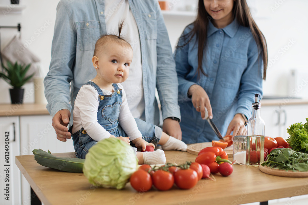 Sticker Close up view of adorable infant sitting on dining table among fresh crunchy products ready for garden salad. Cute baby daughter participating in family dinner preparation on sunny day at home.