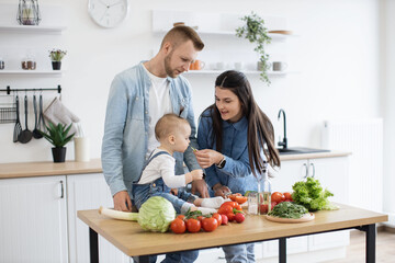 Caring young woman in denim clothes letting baby girl taste green vegetable while cooking dishes for dinner at noon. Mindful parents getting child used to new textures and new flavours of solid food.