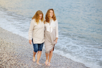 Redhead senior mother and her adult beautiful pregnant daughter are walking together on the sea shore.