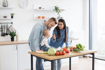 Funny little girl in cozy outfit sitting on dining table while caring young mother holding bite-sized slice of tomato. Mindful parents motivating infant daughter to making healthier food choices.