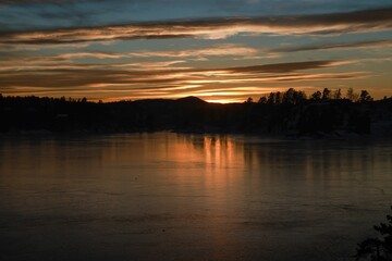 Glowing sun behind the scattered cloudscape over a tranquil lake surrounded by trees at sunset