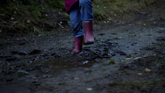 Closeup view of child's feet in pink boots and raincoat, blue jeans jumping into a puddle of mud