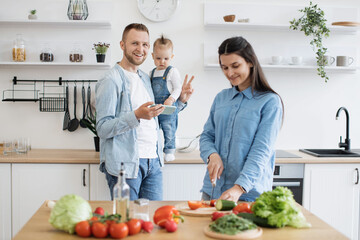 Smiling blond man using cell phone while standing near countertop with infant girl in home kitchen. Loving mother utilizing clean raw ingredients for homemade dishes to please entire family.