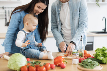 Close up view of male cutting raw vegetable while cute little lady gazing at father's fingers with knife. Adorable baby daughter following the process of food preparation on breakfast table.