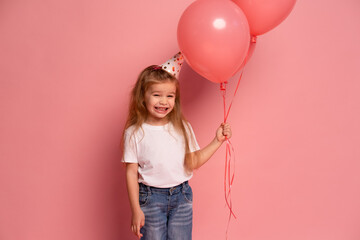 little girl in a white t-shirt and birthday hat holding balloons on pink background