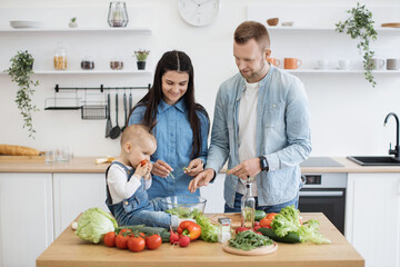 Amusing little child in denim clothes savouring juicy tomato while sitting on table with organic products in kitchen. Cheerful parents feeling excited about new food choice made by baby daughter.