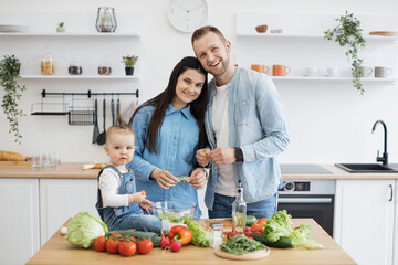 Portrait of joyful caucasian family of three posing in modern kitchen during cooking meals for breakfast. Curious daughter resting near salad bowl while smiling parents snuggling in dining room.