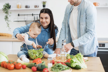 Cropped view of male in denim clothes leaning on wooden table covered with products and kitchenware. Attractive mother using spoons for salad mixture while active kid checking greens in bowl.