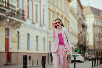 Fashionable elegant confident woman wearing trendy pink sunglasses, suit blazer, satin top, white silk scarf, trousers, walking in street of European city. Copy, empty space for text - 612504427