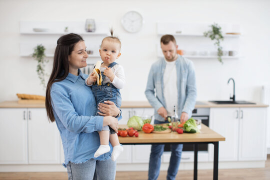 Precious little cutie with ponytail eating organic banana while sitting on mommy's hands. Responsible father cutting and cooking vegetables for lunch on wooden table in white kitchen.