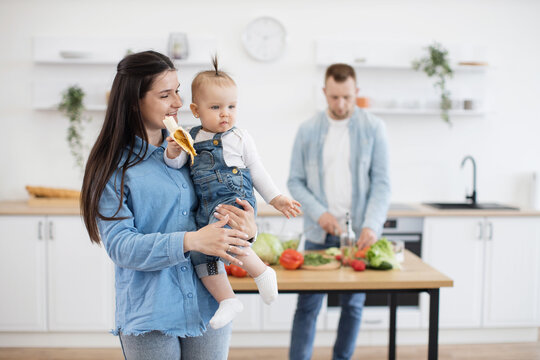 Precious little cutie with ponytail eating organic banana while sitting on mommy's hands. Responsible father cutting and cooking vegetables for lunch on wooden table in white kitchen.