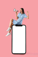 Screaming young woman with megaphone on big mobile phone with blank screen on pink background....