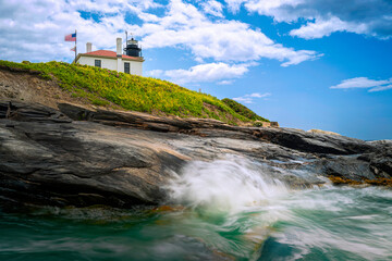 Fototapeta na wymiar lighthouse on the coastal rocks. Summer seascape with splashing misty waves and white clouds in New England