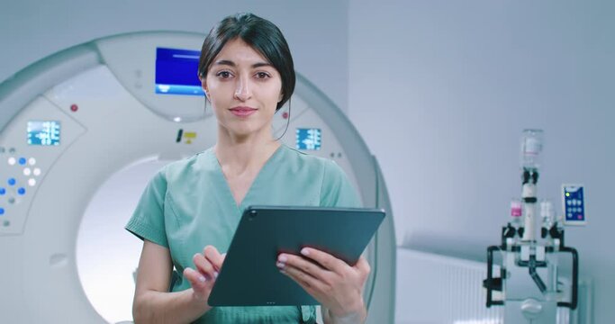 Doctor holds tablet in tomography room. Medical worker in blue coat on background of magnetic resonance capsule. Female doctor is checking scan results. Doctor looks at camera and smiles.