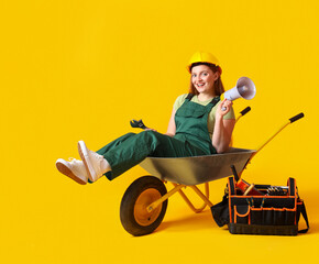 Female builder with megaphone sitting in wheelbarrow on yellow background