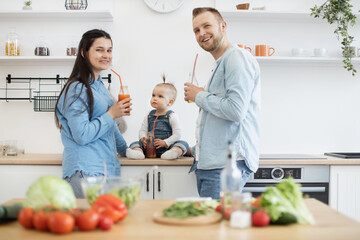 Front view of family trio making healthy meal in kitchen on sunday morning and posing with juice bottles. Happy people wearing denim attire smiling at camera. Concept of home fun and culinary.