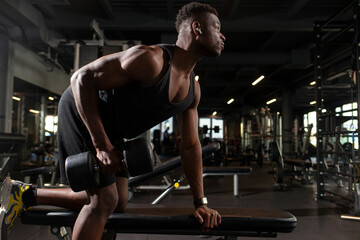 athletic african american man trains in dark gym, athletic guy lifts heavy dumbbells in fitness club