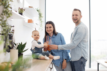 Portrait of two adults and child toasting glasses with fruit juice and smiling at camera. Cute caucasian family enjoying homemade fresh smoothies during healthy diet at home.