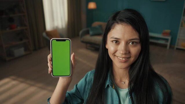 Young smiling woman shows smartphone with green screen. Portrait of an Asian woman with a smartphone in the living room closeup. Technology, gadget, advertising concept.