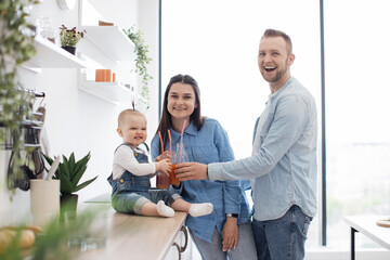 Fototapeta premium Portrait of two adults and child toasting glasses with fruit juice and smiling at camera. Cute caucasian family enjoying homemade fresh smoothies during healthy diet at home.