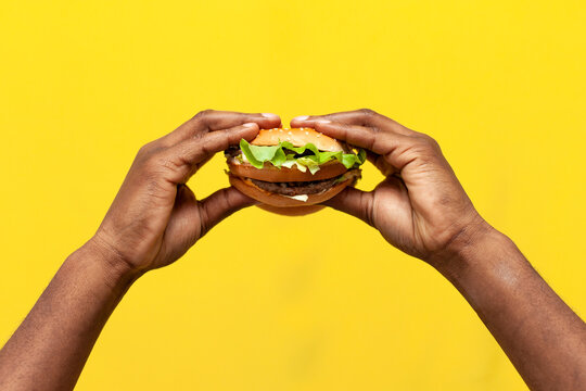 african american male hands hold tasty burger on yellow isolated background, closeup of fast food in the hands