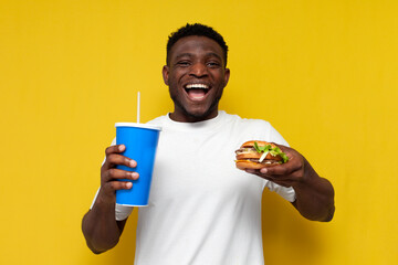 happy african american man in white t-shirt holding burger and soda over yellow isolated background