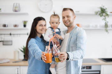 Blurred portrait of caring parents holding little toddler on hands and smiling at camera in kitchen. Cheerful family dressed in casual clothes posing with freshly squeezed juice in bottles.