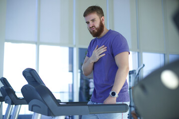 Fototapeta na wymiar Tired exhausted young man is training workout, running on treadmill in gym holding his heart with hand, suffering, feeling unwell, unhealthy during run 
