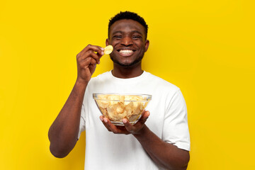 happy african american man holding plate of chips and eating snacks from potatoes on yellow...