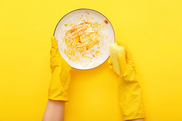 Female hands in rubber gloves washing dirty plate with sponge on yellow background