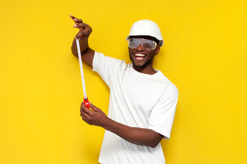 african american male engineer in hard hat and goggles holds tape measure on yellow isolated...