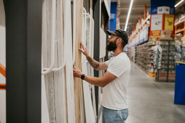 A young handsome man with a beard in casual clothes in a construction hypermarket in the lumber department selects wood building materials for the renovation of his house. - obrazy, fototapety, plakaty